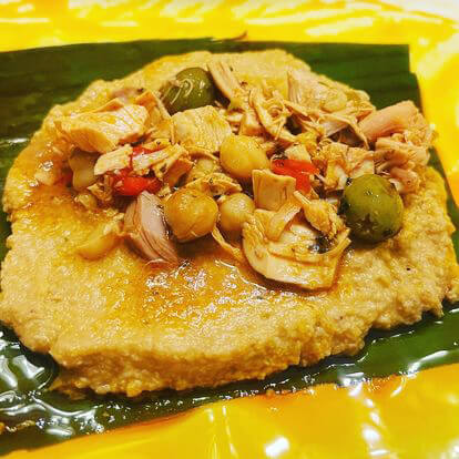 A plate of mashed plantains, topped with fish, beans, and olives on a banana leaf