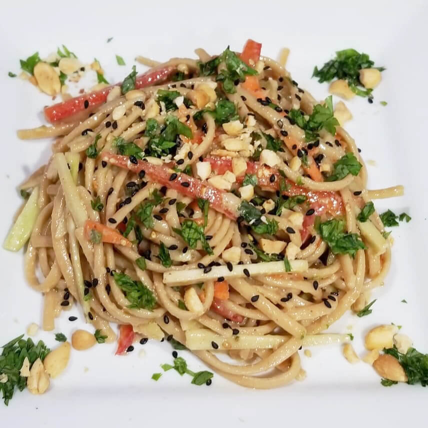 A healthy plate of sesame noodles topped with colorful veggies, nuts, and herbs