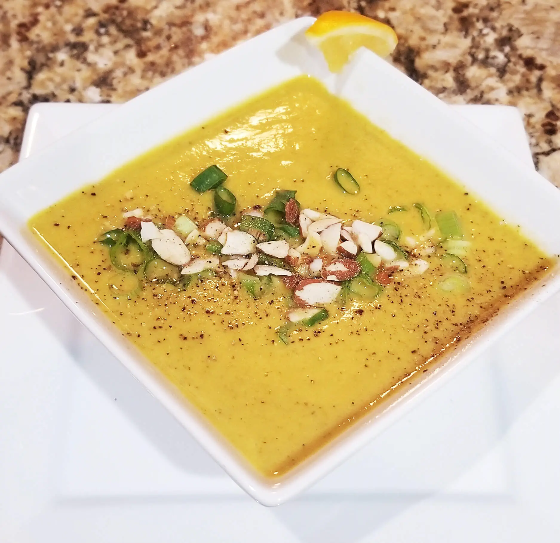 A yellow bowl of soup topped with scallions, almonds and a slice of lemon, offering a delightful blend of flavors and textures