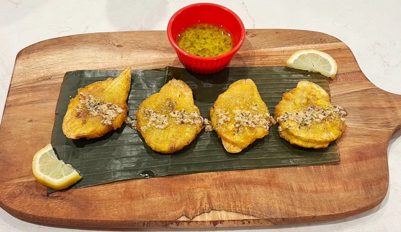 Four crispy snack of fried plantains on a wooden cutting board, with dipping sauce and two lemon wedges